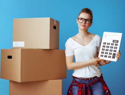 Portrait of pensive modern woman in white t-shirt near cardboard box with calculator against blue background. stay on budget.